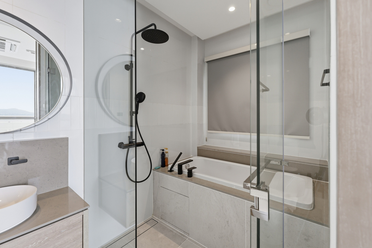 Bathroom with glass-enclosed shower, rainfall showerhead, round mirror, and light wood vanity with white sink.