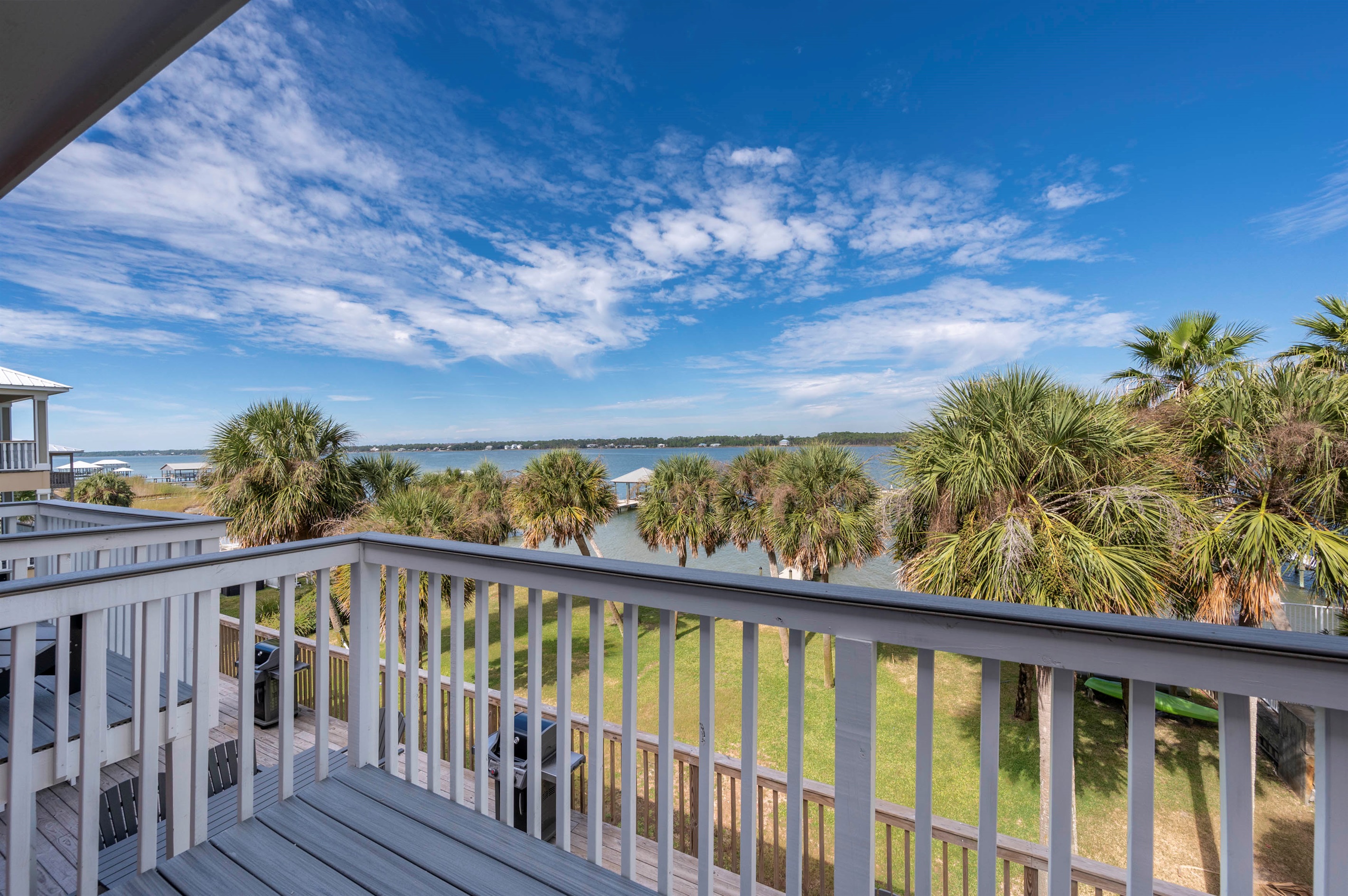 Relax on a balcony at “Gone Coastal” in Gulf Shores, AL, with stunning waterfront views, clear blue skies, and swaying palm trees.
