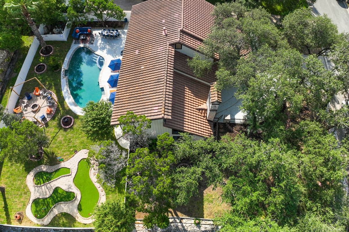 A stunning aerial of The Comfy Coyote. A spacious corner-lot retreat with lush trees framing the front lawn and a resort-style backyard complete with pool, mini golf, hot tub, and firepit.