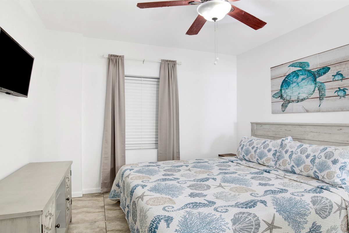 Bedroom with king bed, wall mounted TV, ceiling fan, dresser and ensuite bathroom