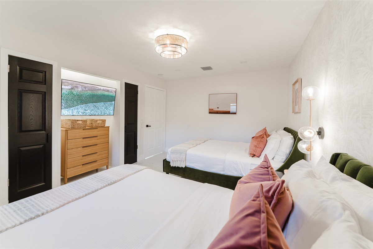 Bright coastal suite with two queen beds, private ensuite bath, smart TV, blackout bamboo shades, and a rattan ceiling fan—ideal for families or shared stays.