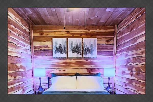 Rest easy in this inviting queen bedroom featuring cedar-lined walls, soft lighting, and forest-inspired decor. A classic buffalo plaid throw and cozy quilt add warmth, while a ceiling fan overhead keeps the room fresh and relaxing all night.