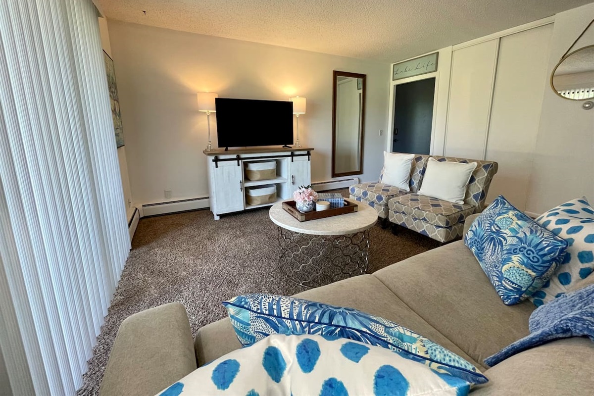 Bright and cozy living space with plush seating, calming blue accents, and a large smart TV for relaxing nights in. Natural light pours in through the sliding glass door, adding warmth to this charming retreat—perfect for comfort and convenience.