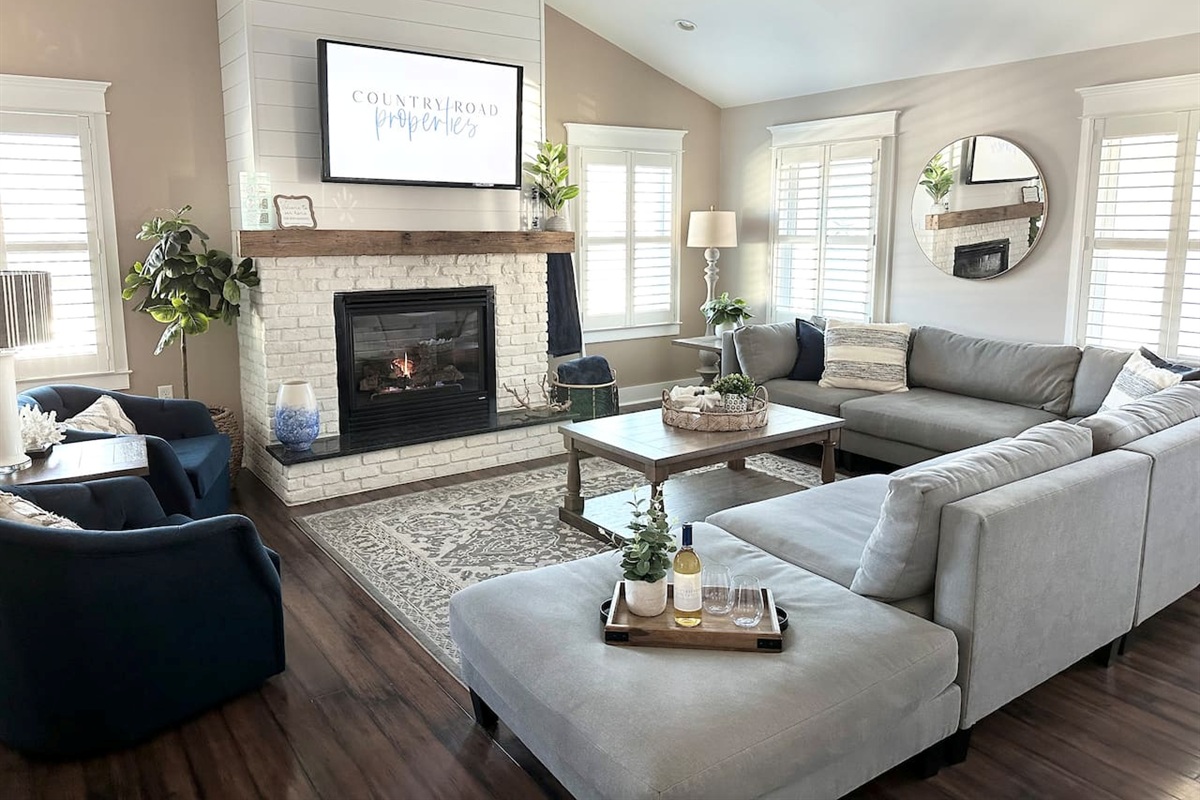 Gather in this sun-drenched living room, featuring vaulted ceilings and a cozy white brick fireplace. The sprawling grey sectional and navy accent chairs offer plenty of space to relax and watch a movie after a long day at the beach.