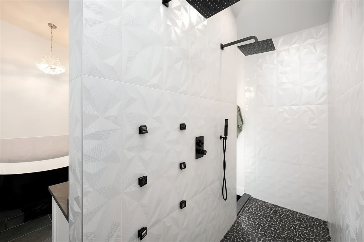 Striking 3D textured wall tiles, a sleek pebble stone floor, and a dual rainfall showerhead system with body jets. A modern, high-end sanctuary designed for a premium spa-like retreat.