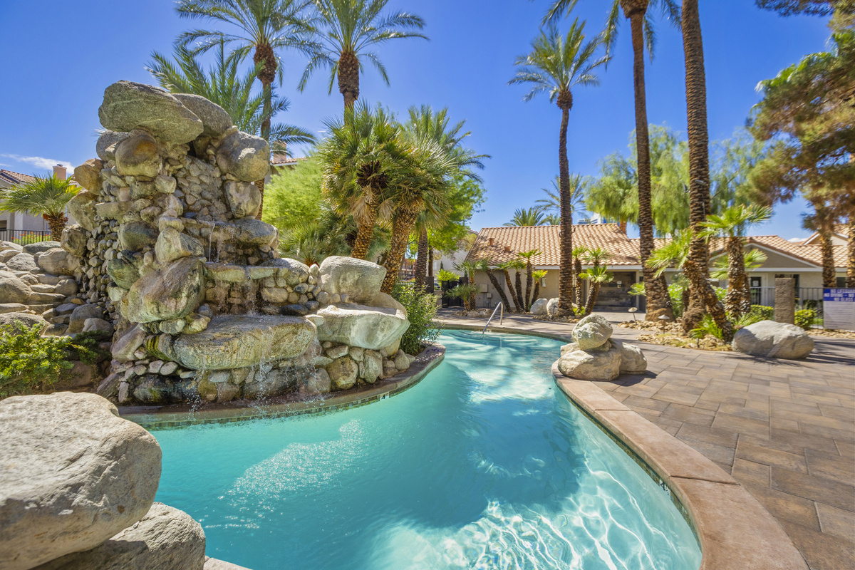 Shared resort-style pool with rock waterfall, palm trees, paved lounge area, and shaded spots around the water.
