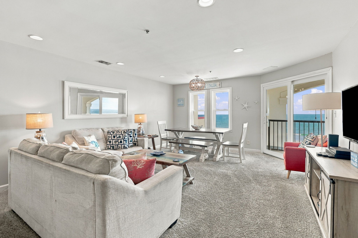 Sunlit, open-concept living space highlighting comfortable sofa seating, a weathered wood coffee table, an adjacent dining setup, and oversized glass doors opening directly to sweeping ocean horizon vistas.   