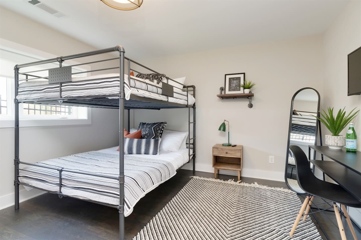 - Metal bunk bed with striped bedding offers a full size lower and twin upper setup - Includes a wood nightstand, reading lamp, and minimalist desk with modern mirror - Bright window and geometric rug enhance the clean, contemporary vibe