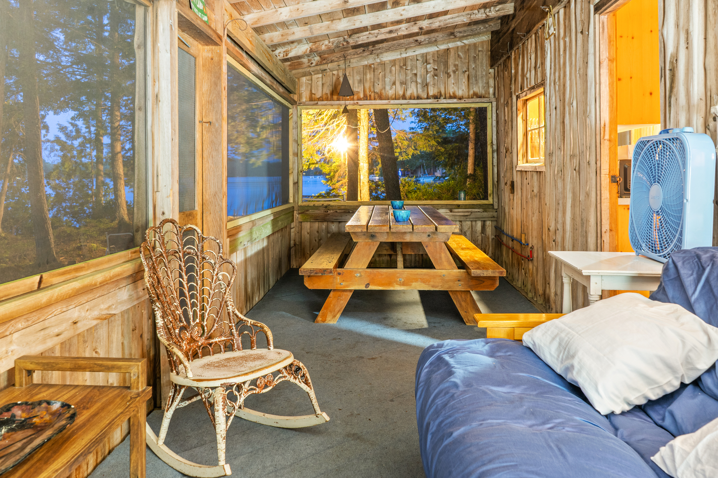 Screened porch dining setup at Loonrise Point! Classic wooden picnic table offers bug-free outdoor meals with sunset lake views. Complemented by wicker rocking chair and versatile futon lounge on Beech Hill Pond in Otis, ME.