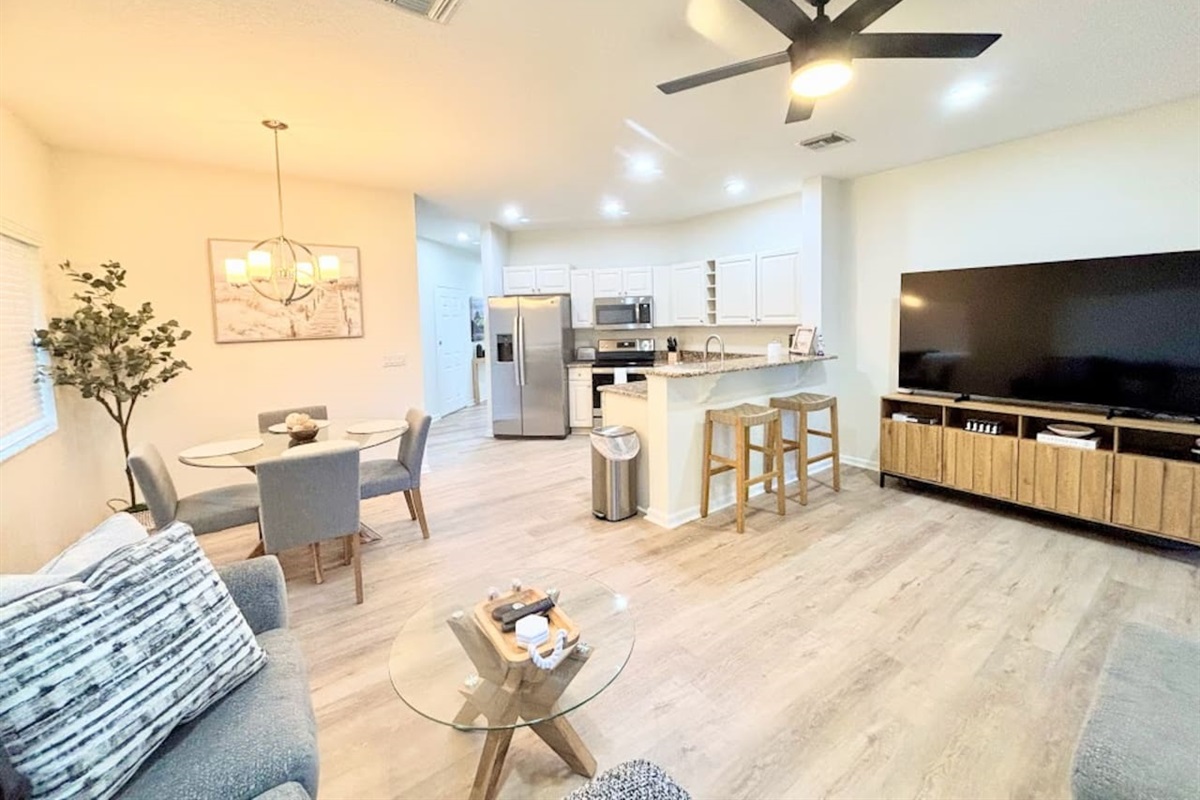 Spacious modern open-concept living area with stylish furnishings, large TV, dining space, updated kitchen, and bright wood-look flooring throughout.