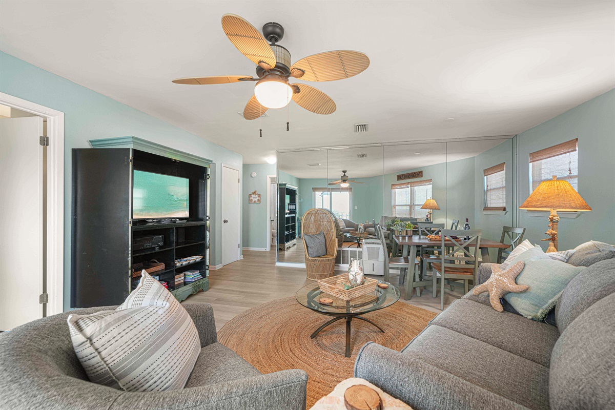 The living room flows into the dining area and out to the deck. Flat-screen TV, wicker peacock chair, and room for all 6 guests.
