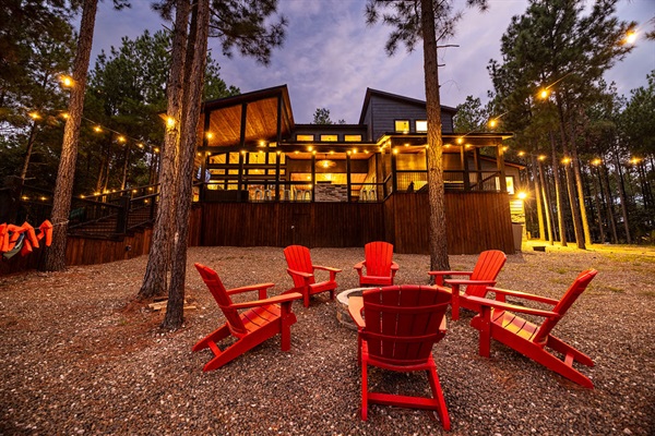 Gather around a cozy fire pit with vibrant red Adirondack chairs, surrounded by towering pine trees and ambient string lights—an ideal setting for evening relaxation at this stunning vacation cabin.