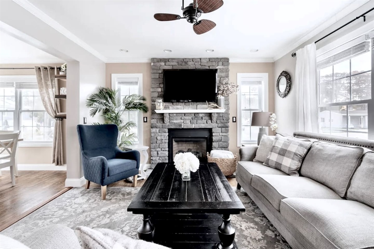Relax in this inviting living space featuring a floor-to-ceiling stone fireplace, a large flat-screen TV, and a cozy sectional. Large windows and a ceiling fan create a bright, comfortable atmosphere for your stay.