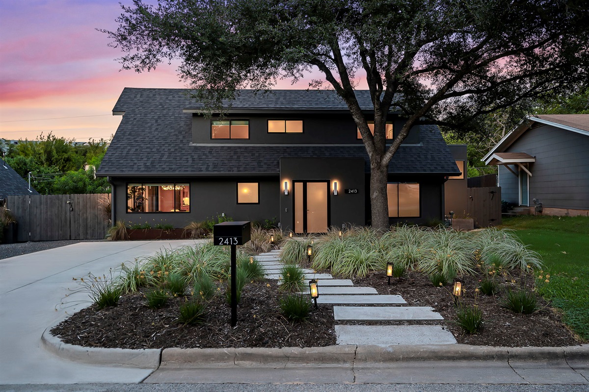 complemented by a beautifully manicured front yard featuring a meandering stone walkway, lush grasses, and accent lighting that creates a welcoming glow at dusk.