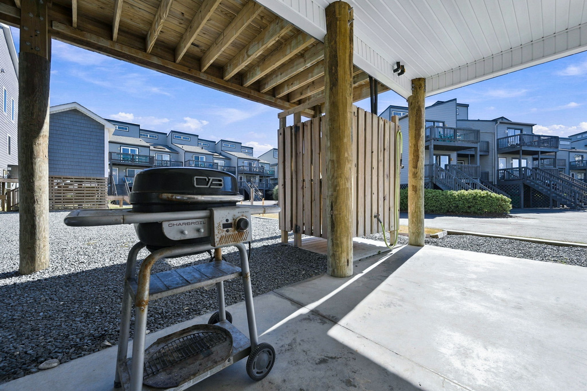 Covered ground-level patio area featuring a outdoor grill and an enclosed wooden outdoor shower under the elevated structure.   