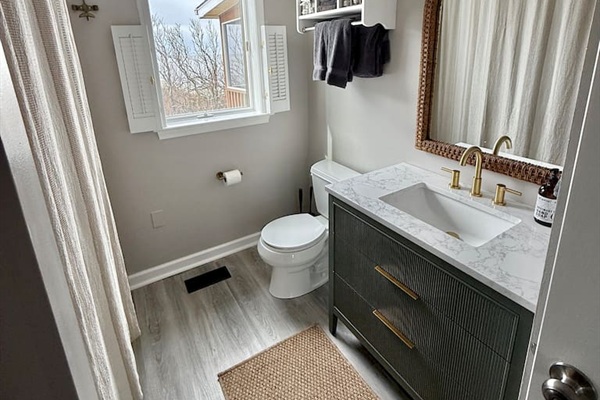 This clean, stylish bathroom features a modern dark vanity with gold hardware and marble countertops, paired with a woven rug and natural light from shuttered windows to create a serene, coastal atmosphere.