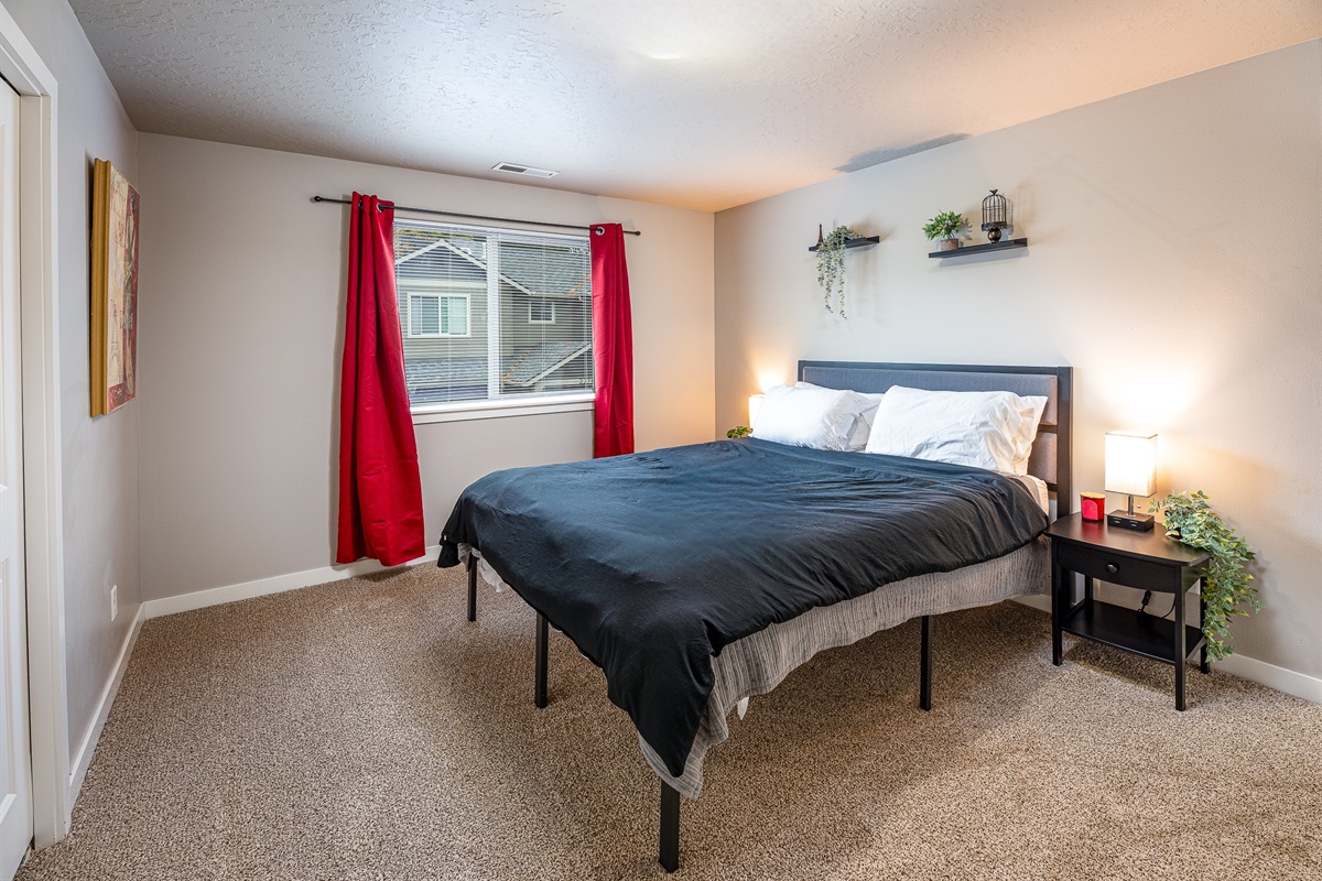 A cozy bedroom featuring a black bedding set on a bed frame, with a nightstand beside it holding a lamp and decorative plant, red curtains framing a window, and artwork on the walls.