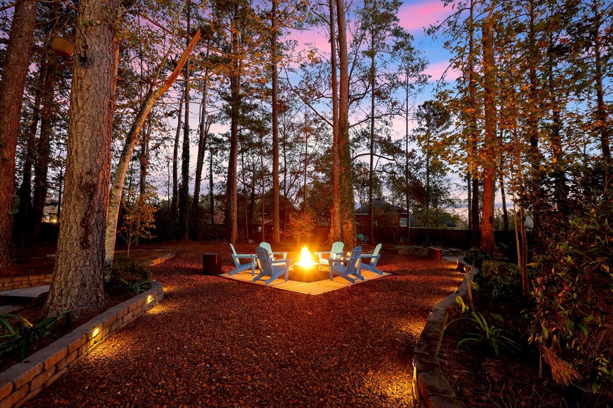 Adirondack chairs around the lit fire pit at dusk under tall pines.