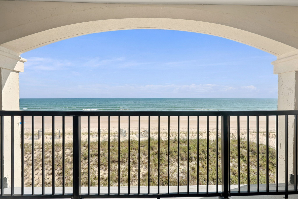 Framed ocean-horizon vistas looking out across natural beach dunes, viewed from behind dark balcony railings beneath an arched stucco overhang.   