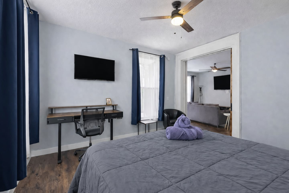 Bedroom workspace angle featuring a desk, ergonomic chair, wall-mounted TV, and natural light.
