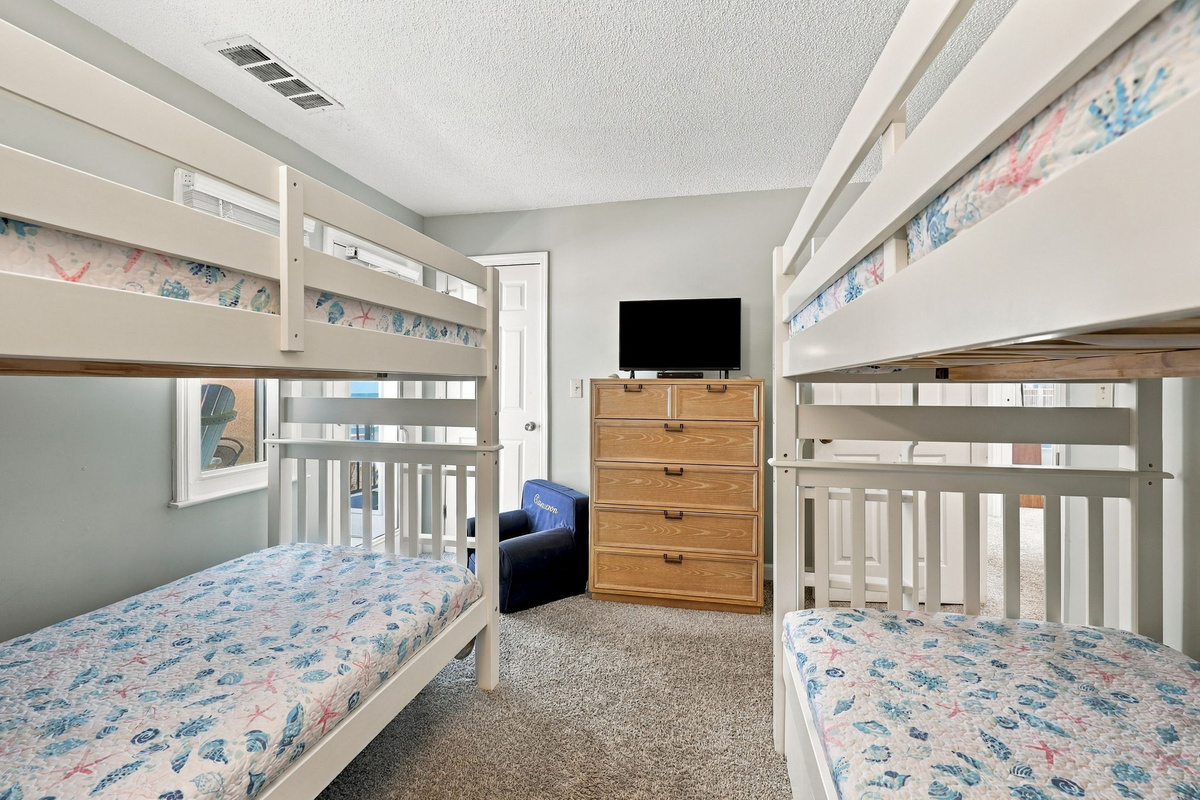 Custom bunk room perspective showcasing two white wooden bunk beds dressed in coastal-chic seashell bedding, framed by soft gray walls, a honey-toned wood chest of drawers with TV, and a cozy navy lounge armchair.   