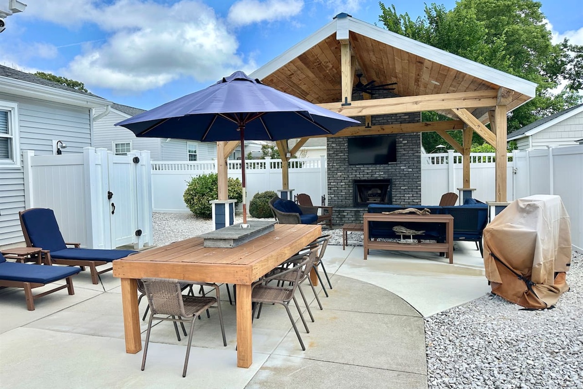 This backyard oasis features a massive wooden pavilion with a stone fireplace and wall-mounted TV. The space is fully equipped for relaxation and dining, including lounge chairs, a dining table with a blue umbrella, and a professional grill.