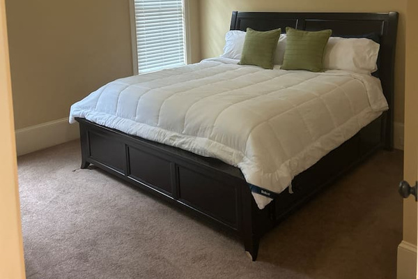 “Bright, comfortable bedroom with a queen bed, plush bedding, and soft neutral tones. Features a large window with blinds, cozy carpet, and ample space to relax—perfect for a restful night’s sleep.”