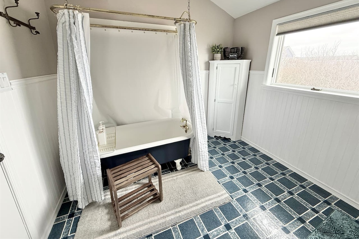 This spacious a coastal-style bathroom is highlighted by a clawfoot tub with a striped shower curtain, a wooden stool, and blue and white checkered tile flooring.