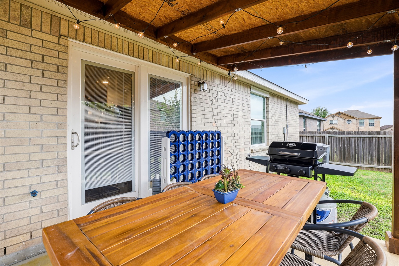 Covered patio with string lights, wood dining table, propane BBQ grill, and lifesize Connect 4 — your own private outdoor entertaining space. Direct access from the kitchen through sliding glass doors.