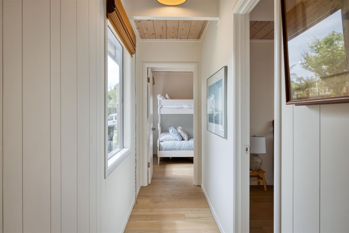 - Hallway with natural light from side windows leading to cozy bunk room  
- White paneled walls and wood ceiling create a fresh coastal inspired feel  
- Artwork adds character to this inviting passageway between bedrooms  
