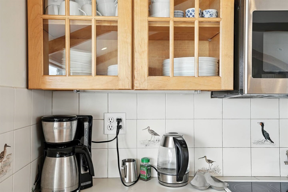 - Coffee station with drip coffee maker, electric kettle, & essential supplies for your morning  
- Glass front cabinets neatly display cups, plates, & bowls for easy access  
- Decorative tile accents with coastal bird designs add charm to the space
