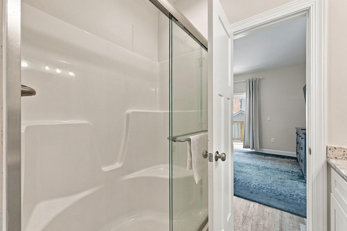 Bathroom view showcasing the glass-enclosed tub/shower combination with molded seating, looking through the doorway into the light-filled third bedroom.   
