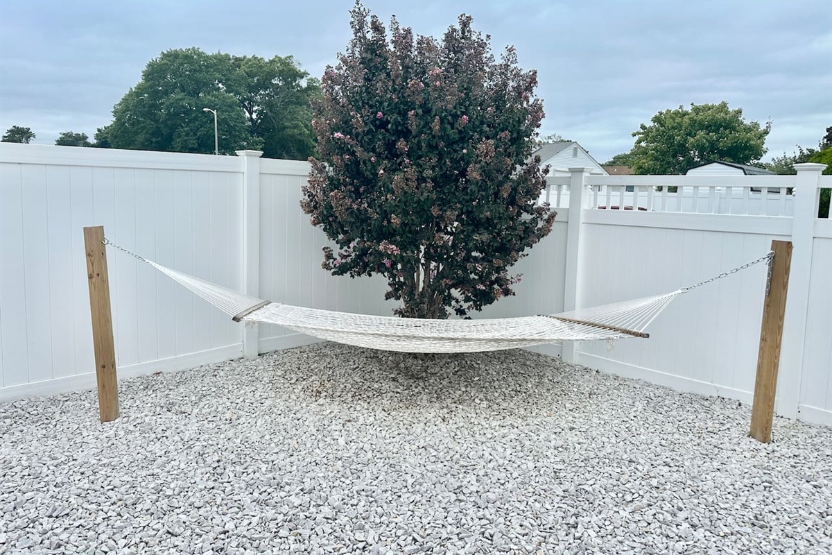 Escape to this peaceful corner of the private, fenced backyard, featuring a comfortable white rope hammock perfect for an afternoon nap. The hammock is set over a clean gravel base, offering a quiet spot to relax in the shade of a lush tree.