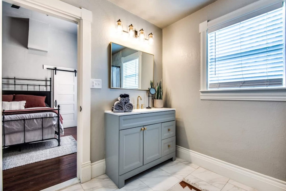 Bright vanity area with sage green double vanity, warm mirror lighting, wood-look tile floors, and a natural light window. Shared between Bedrooms 2 and 3 in this 3BR/2.5BA modern farmhouse Airbnb in downtown Aubrey TX.