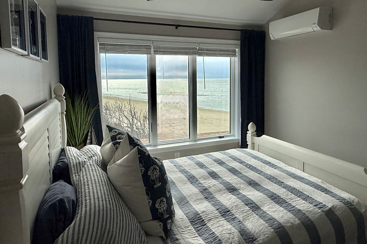This serene bedroom features a white bed frame dressed in blue-striped bedding. The room includes dark curtains framing a window that offers beautiful, direct water views.