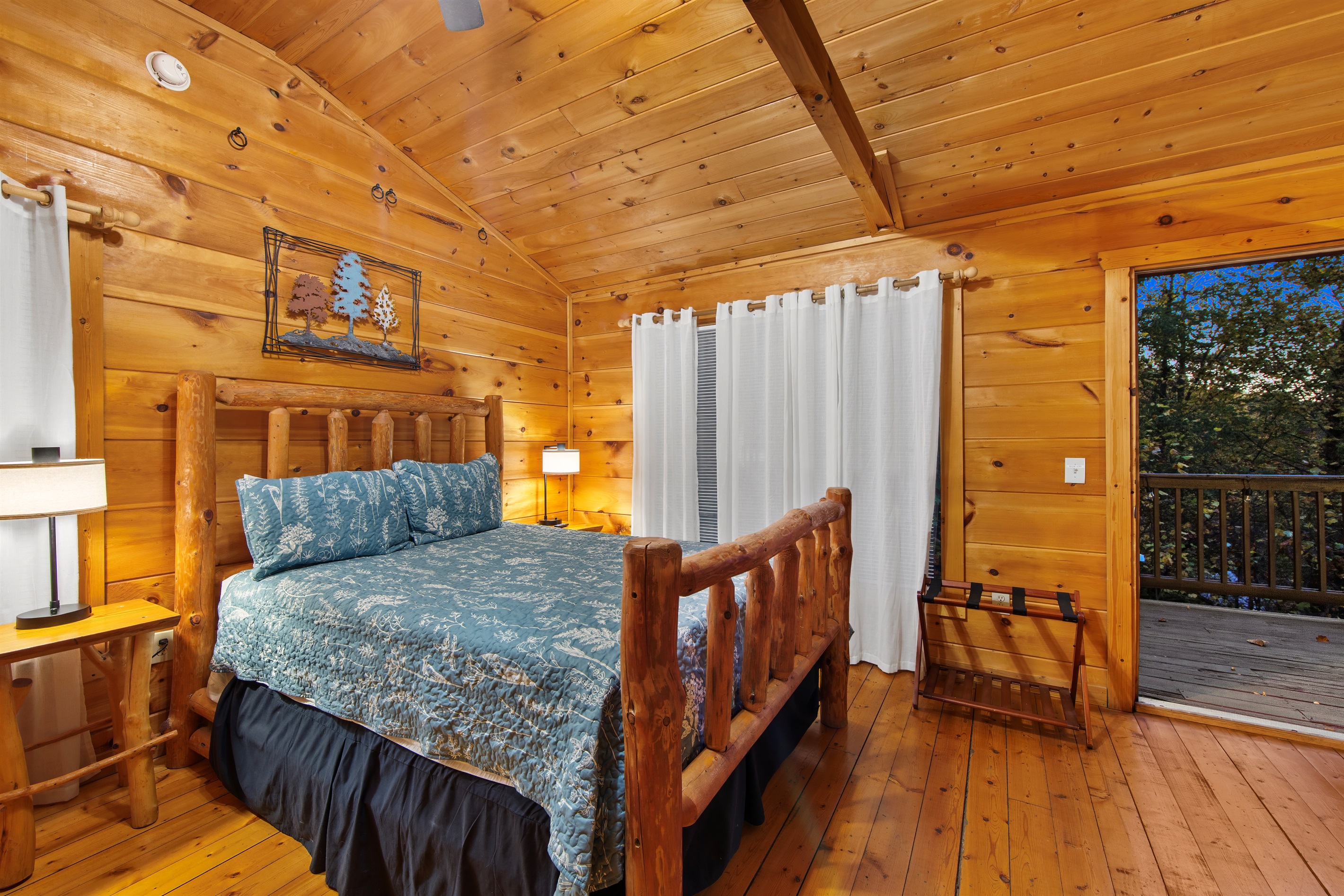 A rustic yet elegant bedroom showcases a log bed with a blue nature-inspired quilt, complemented by warm wood paneling, soft lighting, and a sliding door leading to an outdoor deck.