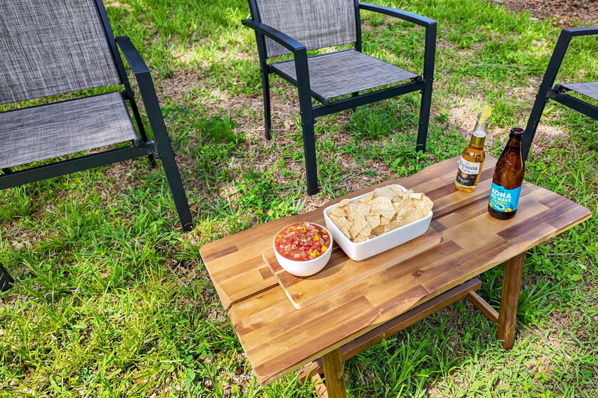 Four modern black patio chairs circle a small wood coffee table, styled with snacks, drinks, and a casual vibe.