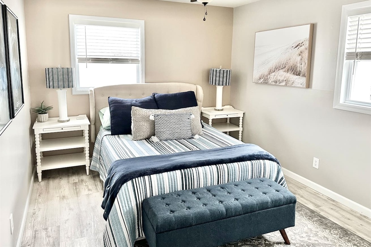 Relax in this stylish guest bedroom featuring a plush bed with nautical striped bedding and a tufted navy bench. With twin nightstands, coastal artwork, and plenty of natural light, it’s a bright and cozy retreat for your stay.