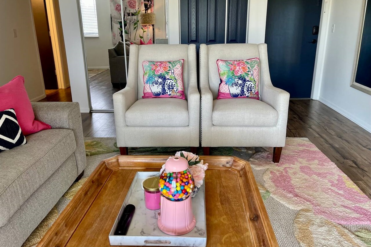 Bright and cheerful seating area featuring two comfy accent chairs with colorful floral pillows. A stylish wooden coffee table adds charm, topped with fun and welcoming decor—perfect for enjoying a cozy, relaxing stay with personality and warmth.