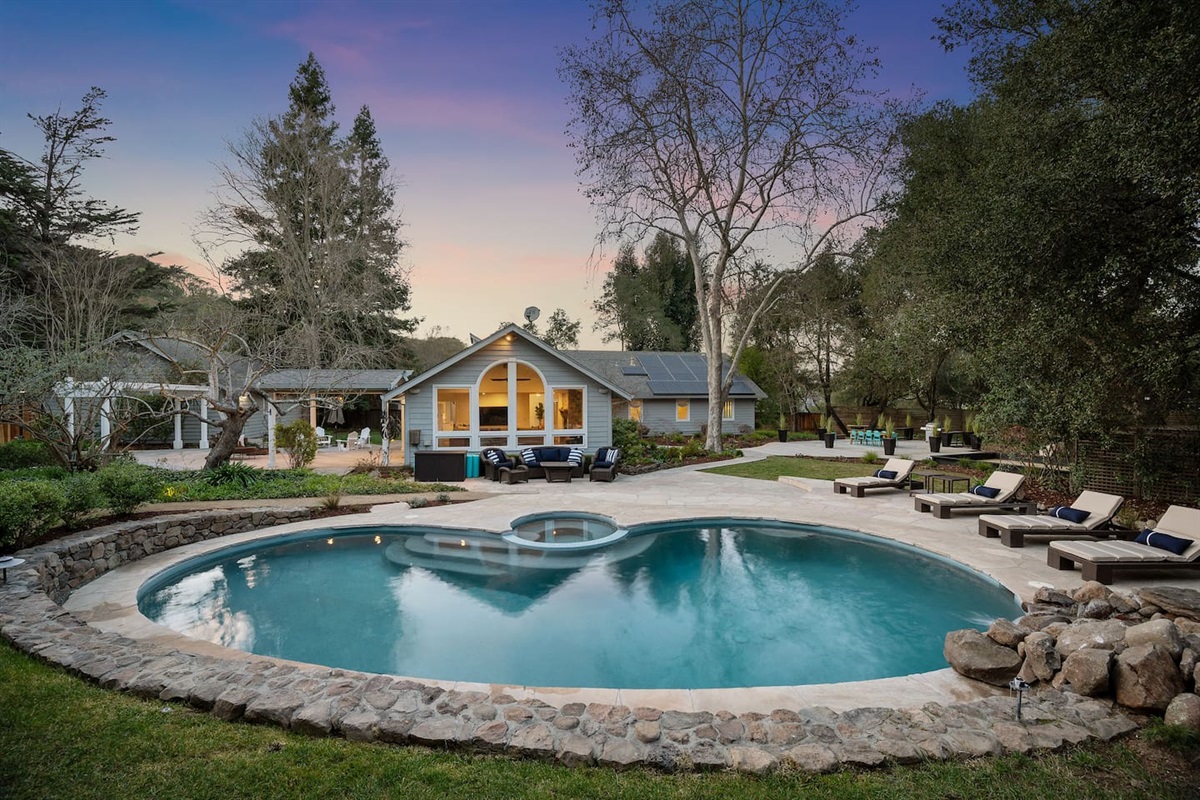To the left, a pool table and seating area outside the kitchen and living room doors. In front, a pool, spa, rock waterfall, pool loungers, and couch + accent chair relaxation space. To the right, the dining pavilion and bocce court.