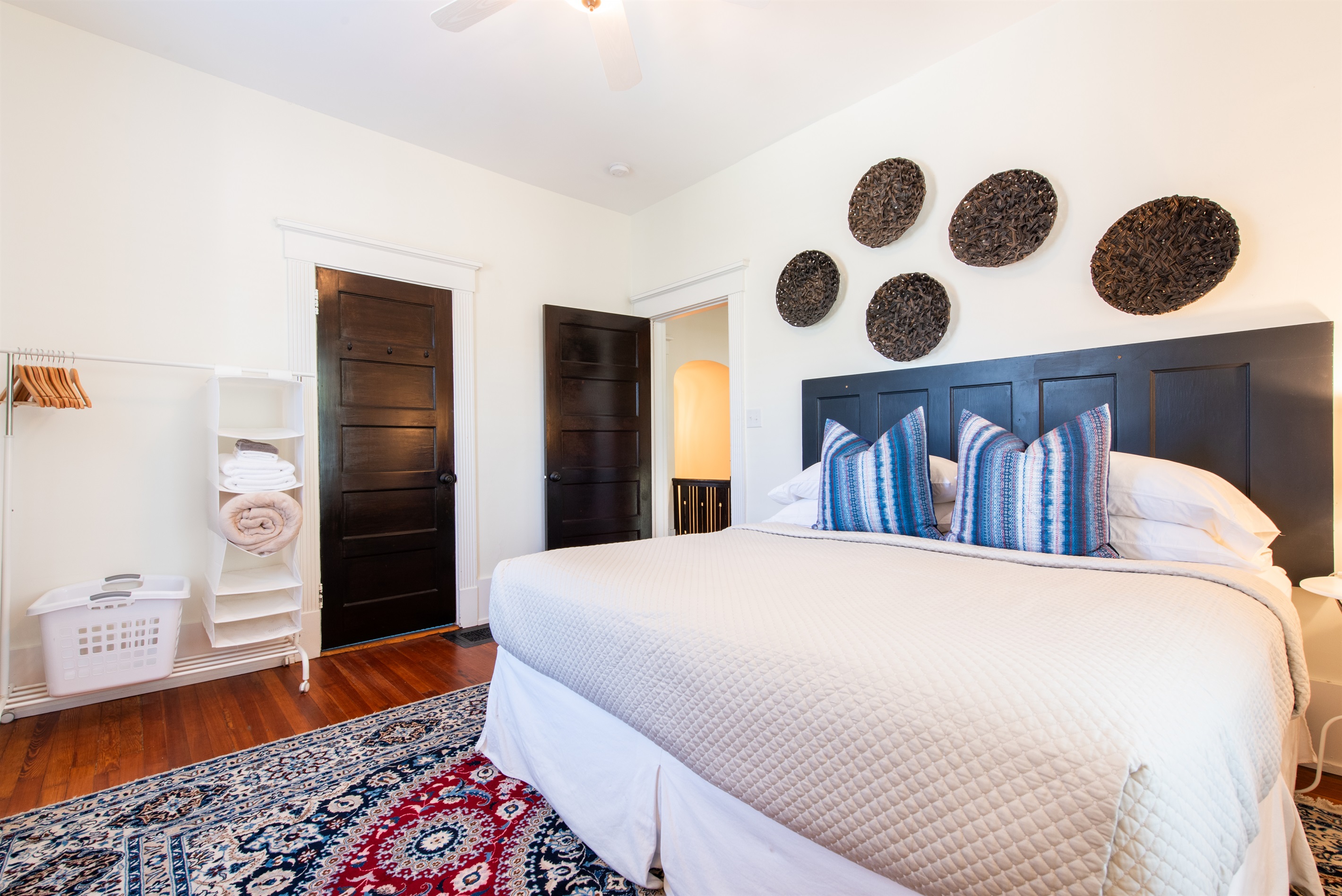 Unwind in this serene bedroom on the Bourbon Trail in Louisville’s Highlands! Features a comfy bed with blue pillows, woven wall art, and a laundry rack—perfect for a restful stay.