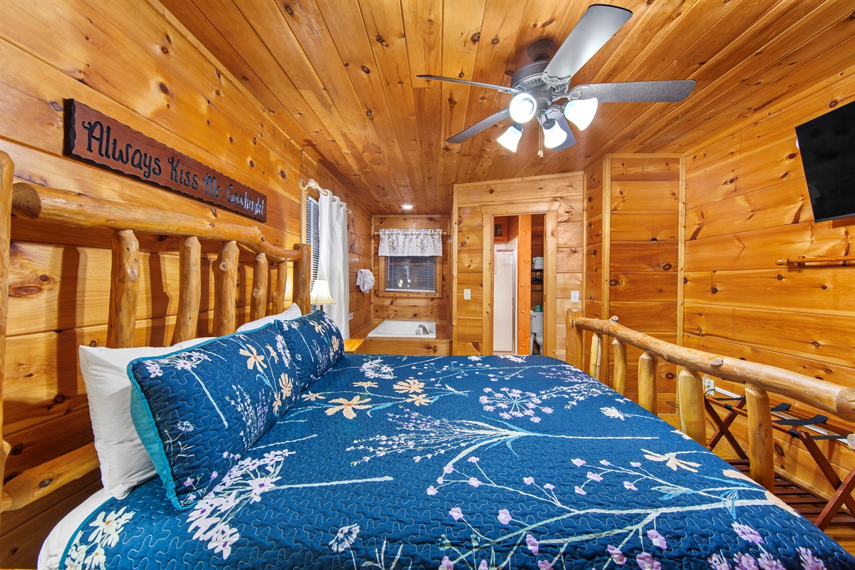 This inviting cabin bedroom showcases a log bed adorned with nature-inspired bedding, soft white curtains, and a jetted tub nearby, offering a perfect blend of comfort and rustic charm.