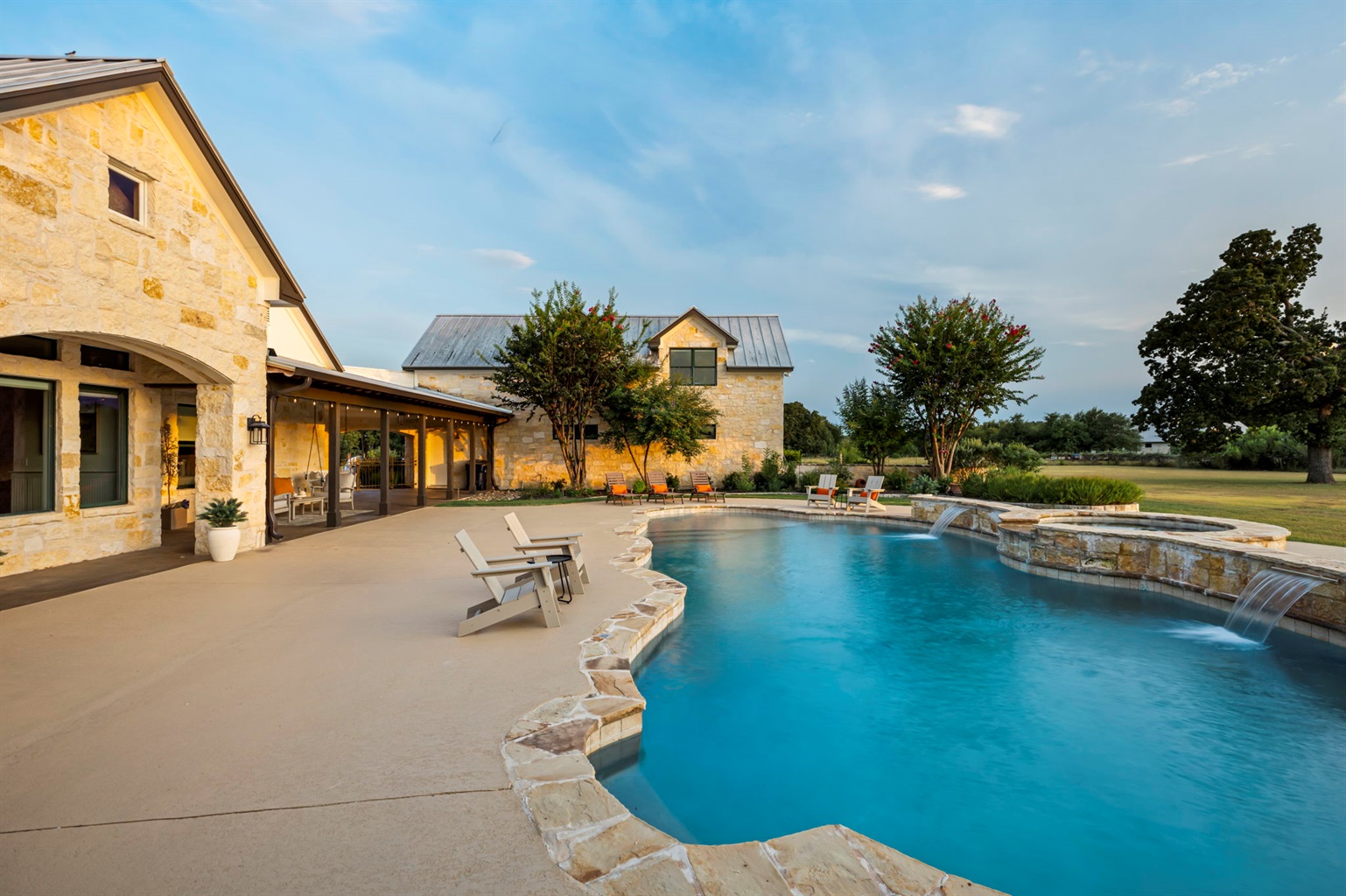 A luxurious Hill Country home featuring a sprawling outdoor living area with a resort-style pool, hot tub, and ample lounging space, set against a clear blue sky.