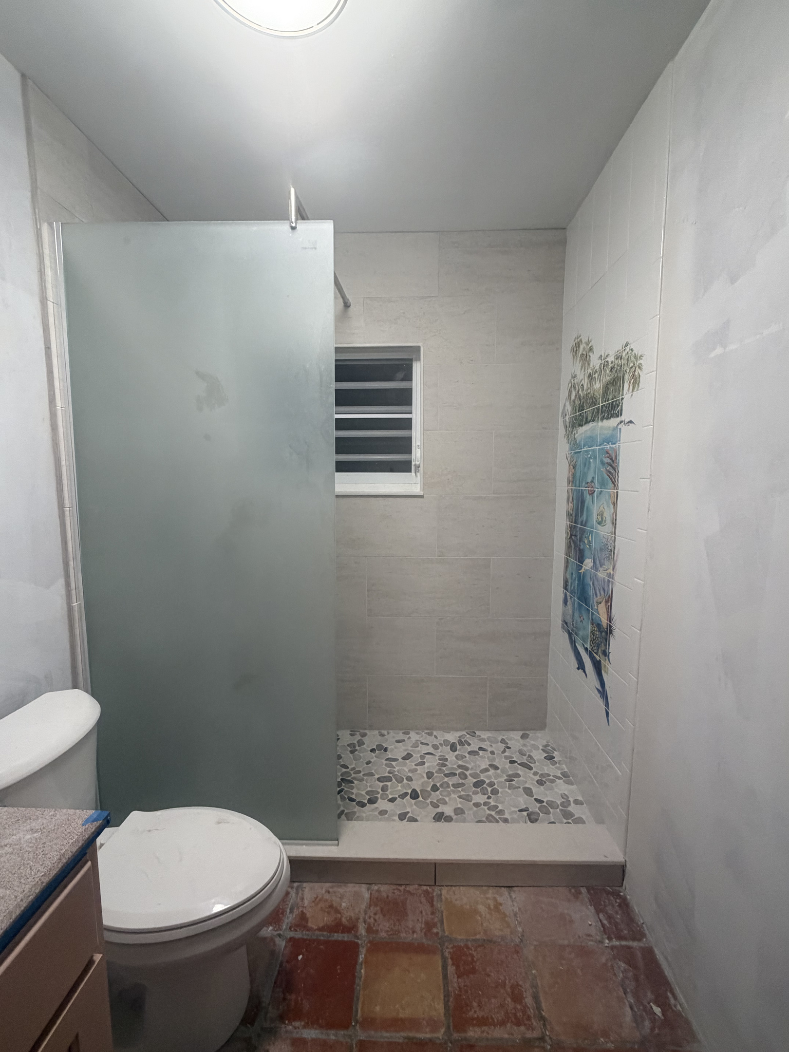 Features a spacious walk-in shower with natural river pebble flooring, frosted glass privacy panel, and USVI artist tile mural featuring dolphins and sea life. Outfitted with a rustic wood-framed vanity mirror, and modern sparkle light fixture. 