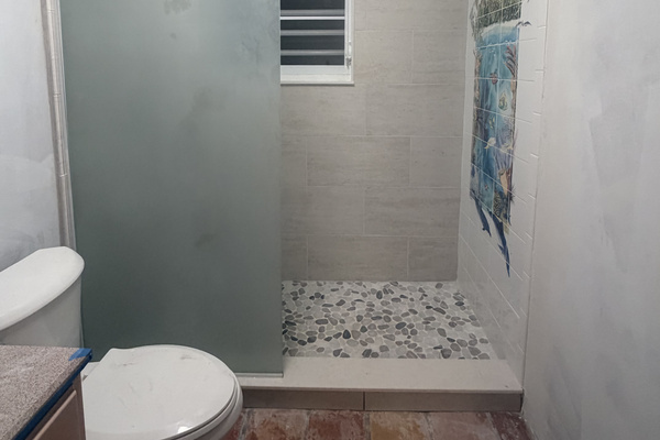 Features a spacious walk-in shower with natural river pebble flooring, frosted glass privacy panel, and USVI artist tile mural featuring dolphins and sea life. Outfitted with a rustic wood-framed vanity mirror, and modern sparkle light fixture. 