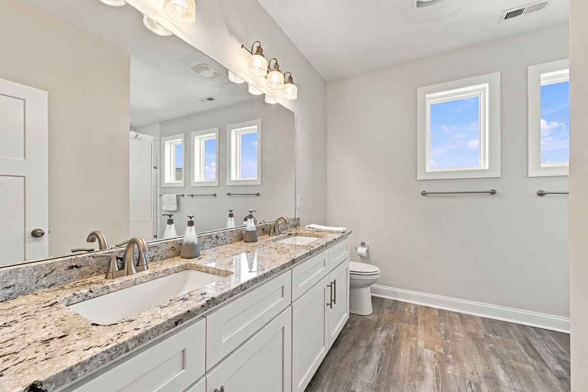 Spacious ensuite bathroom featuring a white double-sink vanity with granite countertops, large mirror, wood-look flooring, and bright accent windows.   