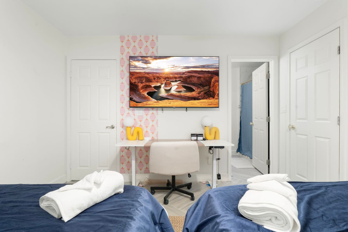 Guest bedroom with dual beds, pink wallpaper, desk, and desert landscape art—comfortable space for couples or shared stays.