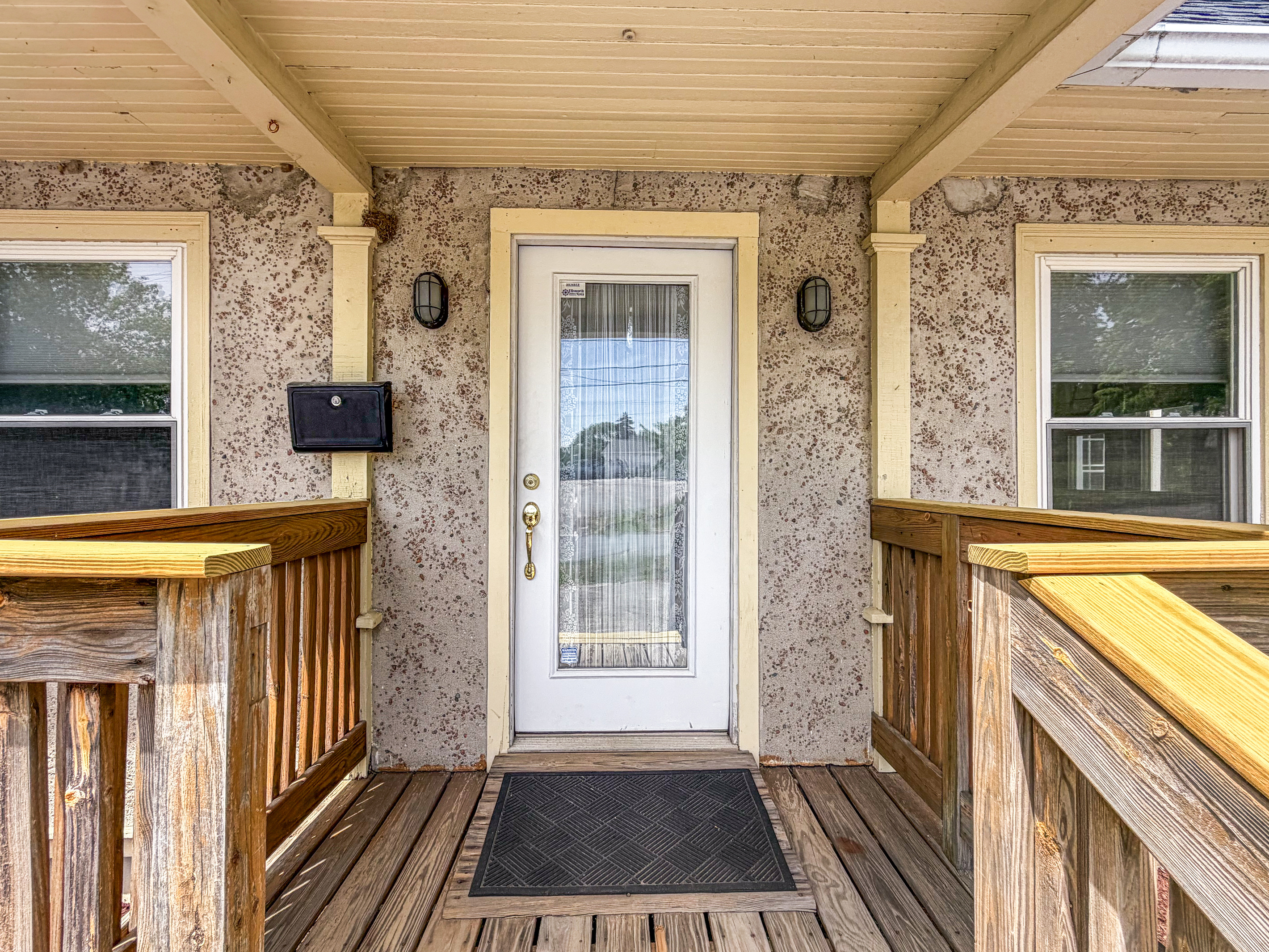 The covered porch deck provides a close-up look at the unique stone-pressed stucco siding and outdoor lighting. While this door forms the classic facade, the active keyless entry pad for guest check-in is located at the side mudroom door.