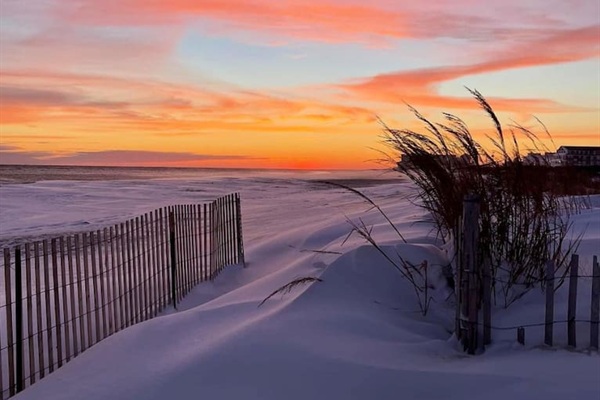 A serene winter scene unfolds as a vibrant, fiery sunset glows over a snow-covered beach. The soft dunes, dusted in pristine white, are lined by a rustic wooden fence, creating a peaceful and picturesque coastal landscape.