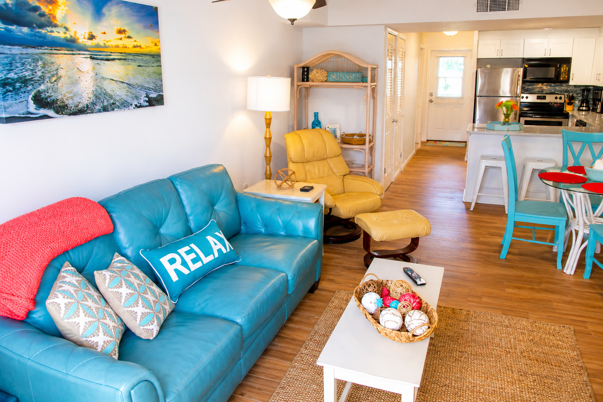 Coastal living room with turquoise sofa, vibrant accents, and modern kitchen for a relaxing stay.
